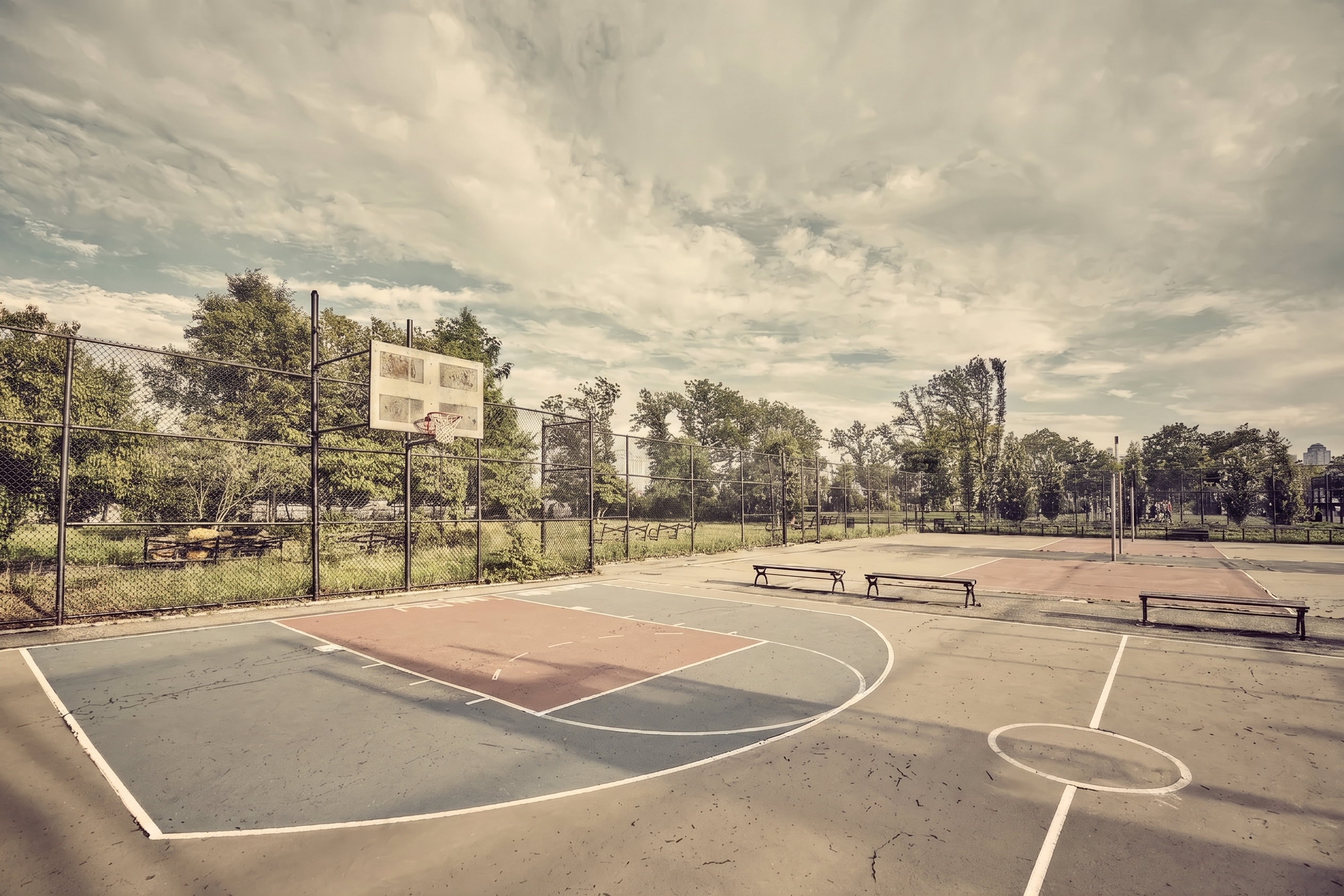 An empty outdoor basketball court enclosed by chain-link fencing and trees.